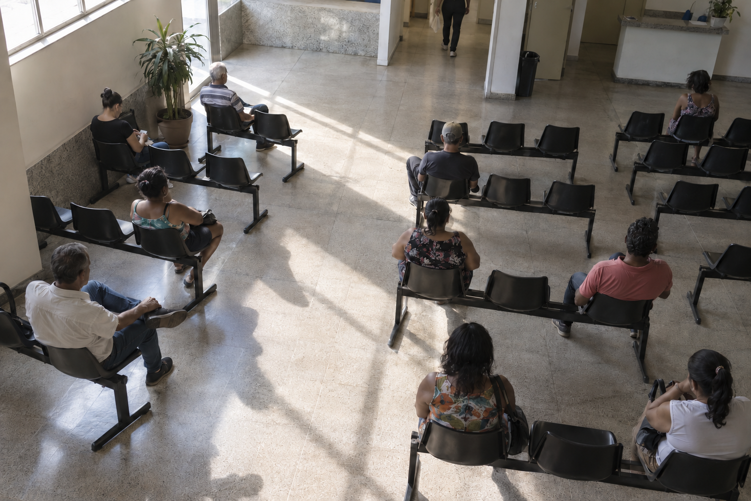 Sala de espera de ambulatório vista de cima, com fileiras de cadeiras plásticas parcialmente ocupadas e cadeiras vazias entre as pessoas.